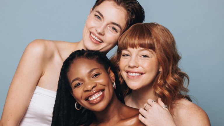 Tri žene različitih tonova kože poziraju nasmejane ispred plave pozadine - slika oslikava značaj melanina u nezi kože