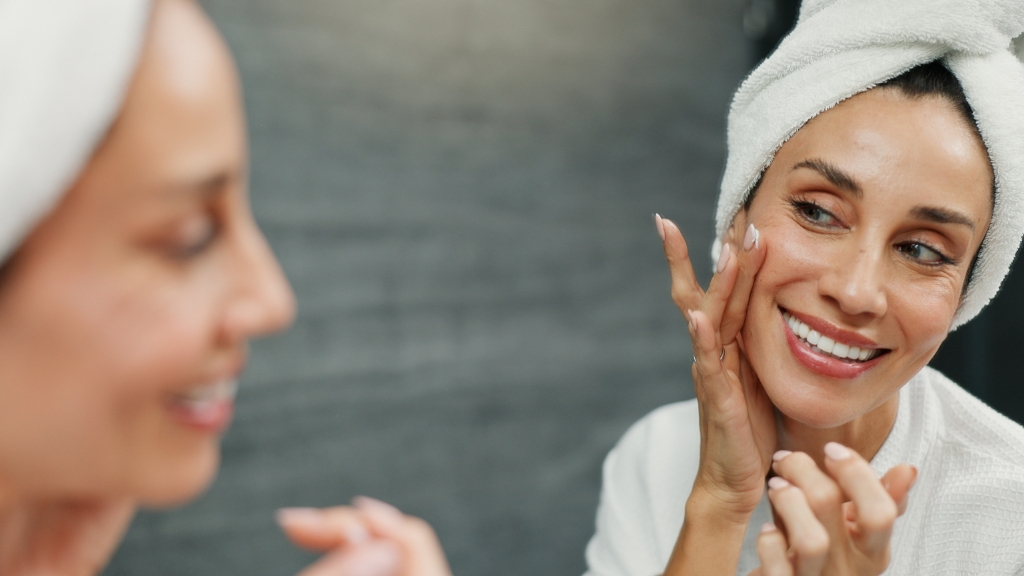 Žena sa peškirom na glavi nanosi kremu na lice ispred ogledala