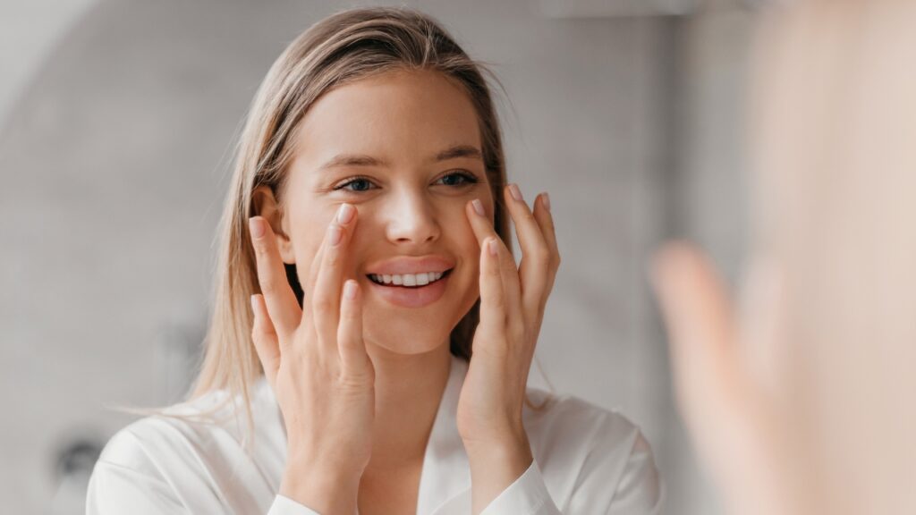 Žena nanosi preparat na lice i proverava u ogledalu kako joj koža izgleda svežije i zategnutije
