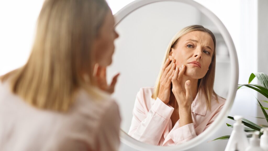 Žena ispred ogledala posmatra lice i primećuje promene na koži dok dodiruje obraz